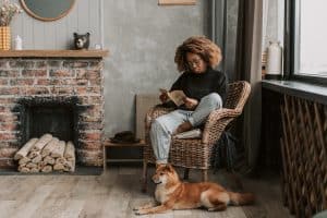 African woman reading a book on a chair at a fireplace with dog lying on the floor